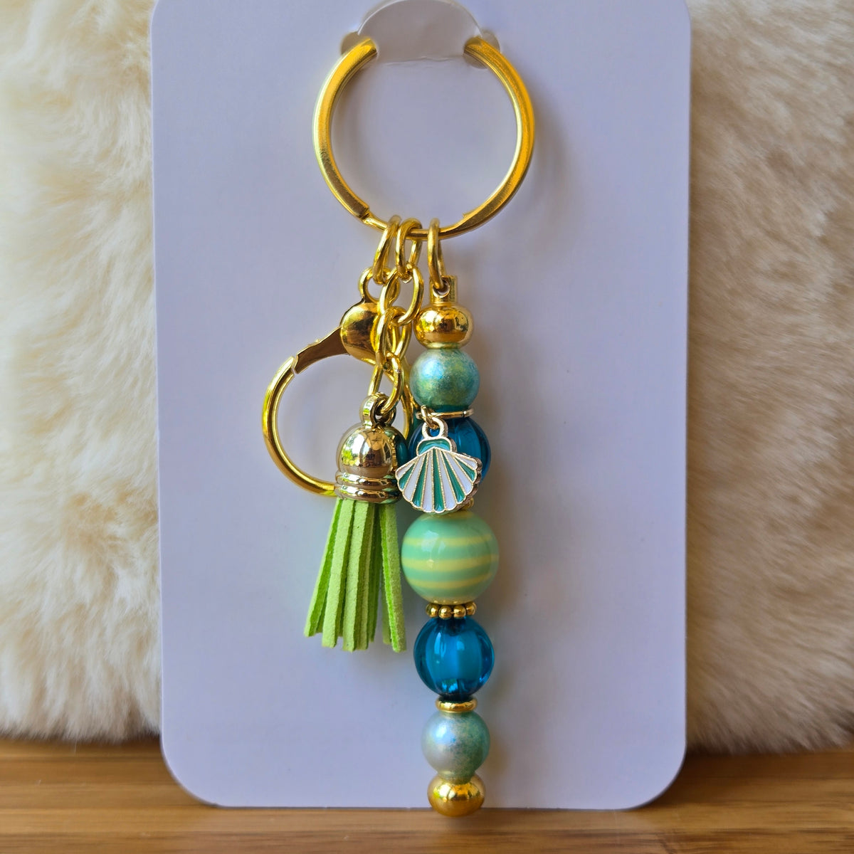 A gold-tone keychain with multiple charms, including a green tassel, blue and green beads, a seashell charm, and a split key ring, displayed on a white card against a soft beige faux fur background.
