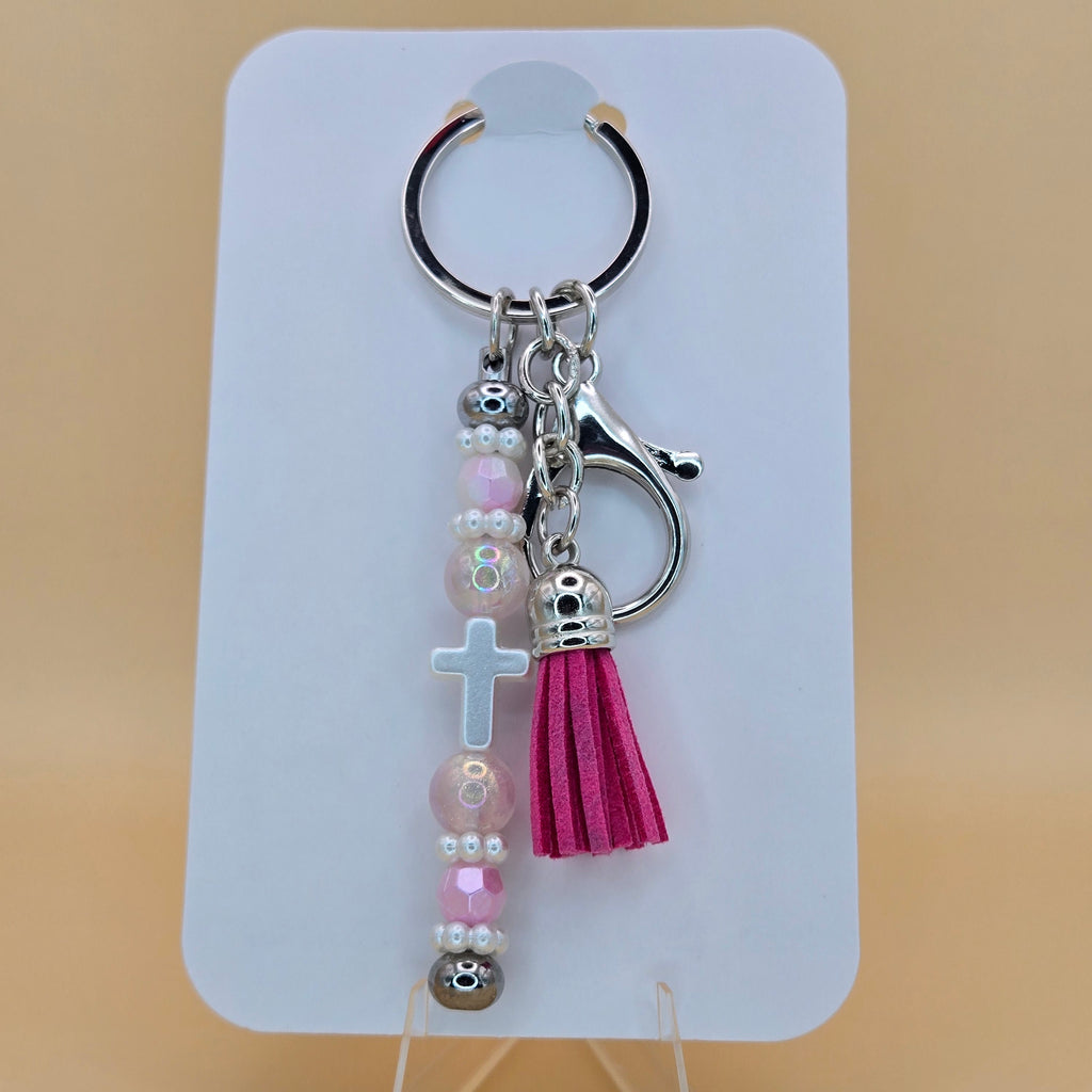A pink and silver beaded keychain with a tassel, skull charms, and a white cross, displayed on a white card stand against a beige background.