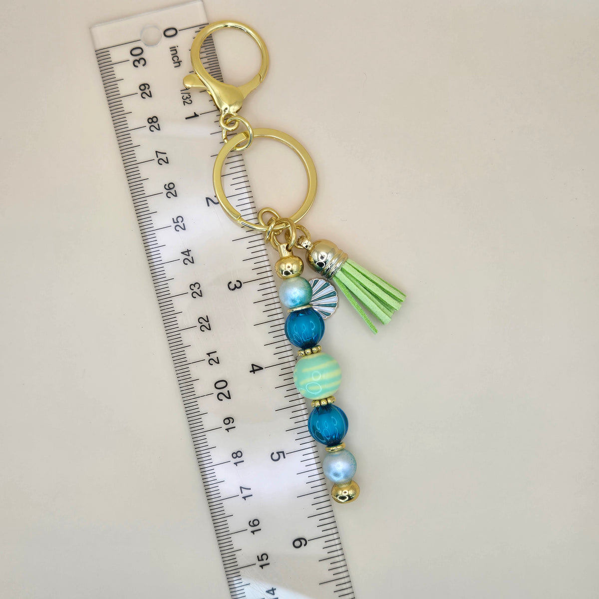 A gold-tone keychain with blue and mint green beads, a striped tassel, and a green paper fan charm, shown next to a clear plastic ruler for scale on a light beige background.