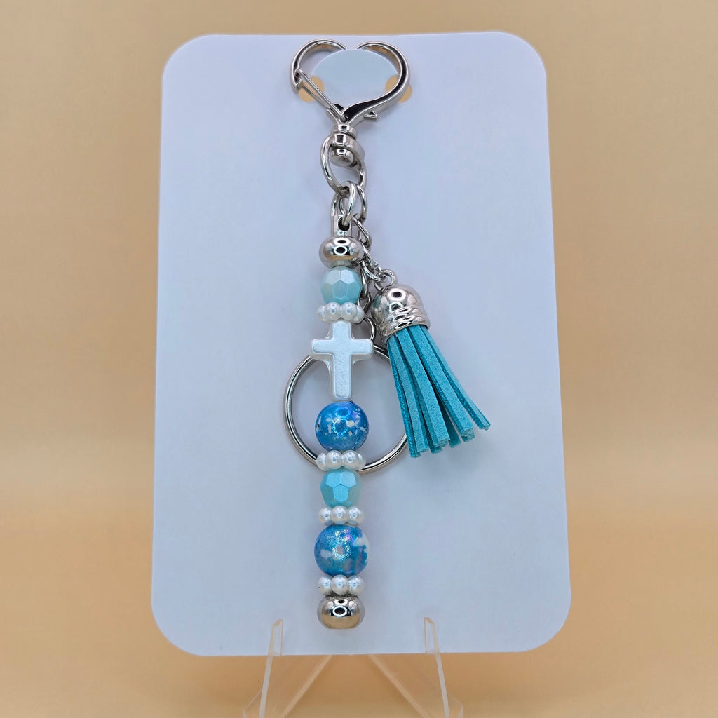 A beaded keychain with a turquoise tassel, silver-toned metal hardware, a white cross charm, and blue marbled beads, displayed on a white card stand against a beige background.